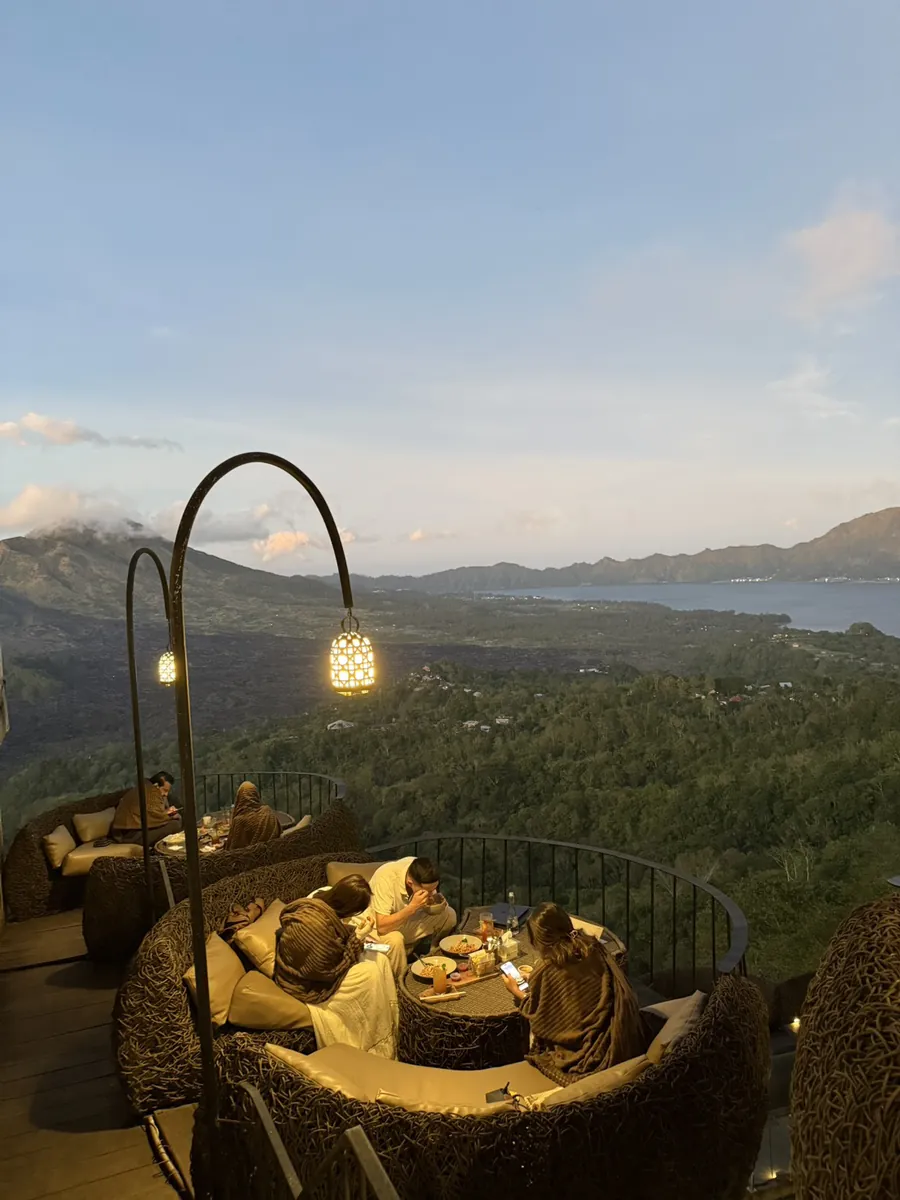 Guests relaxing in woven pod seats under glowing lanterns on the Akasa terrace at golden hour, with Mount Batur and the caldera lake behind, Kintamani, Bali