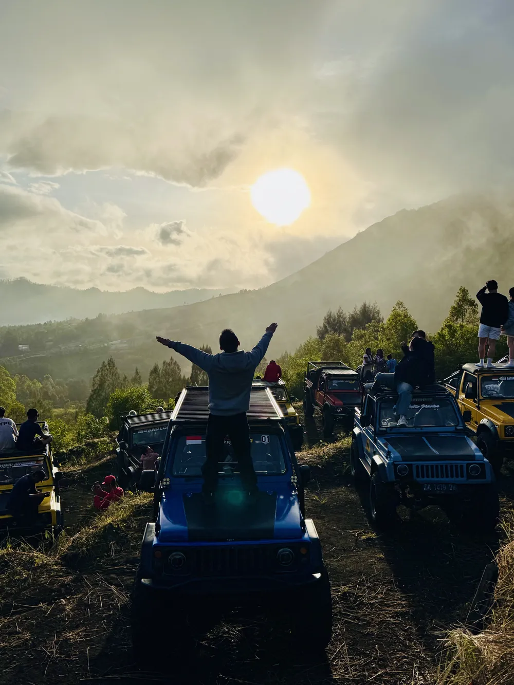 A visitor standing on a black 4WD jeep on the Mount Batur lava field, arms raised, as the sun rises behind the volcano