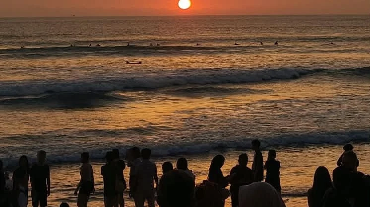 A line of people standing in silhouette along the shoreline at Kuta Beach watching the sun drop to the horizon, surfers waiting in the line-up beyond the shore break, Bali