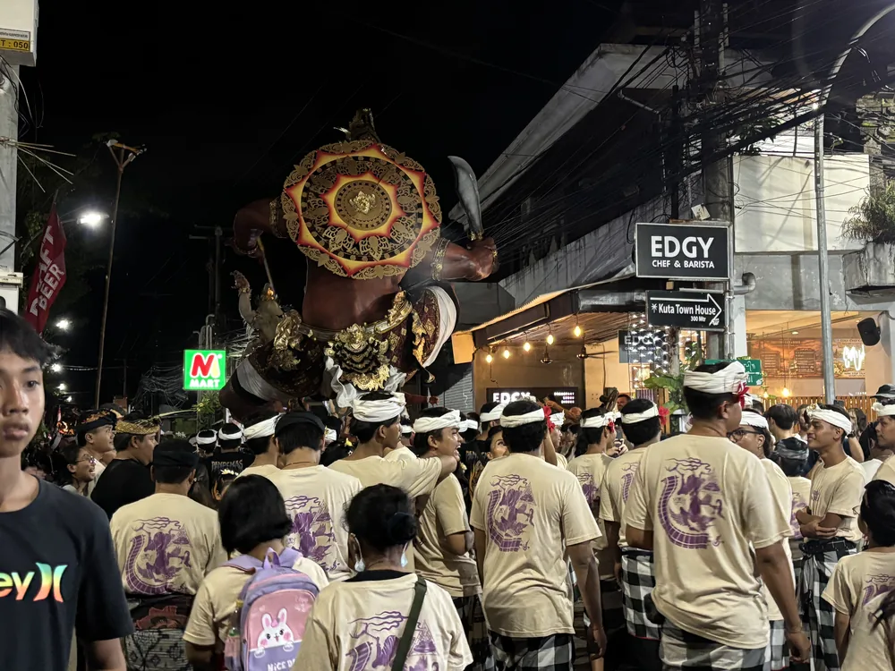 A team in matching shirts carrying an Ogoh-Ogoh down a main street in Kuta with shops and crowds on Nyepi eve, Bali