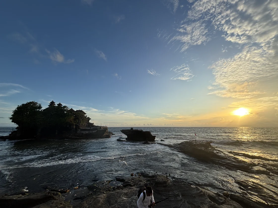 The Tanah Lot sea temple silhouetted at sunset with the tide out, the rock shelf in front of it exposed and the sun low over the Indian Ocean, Bali