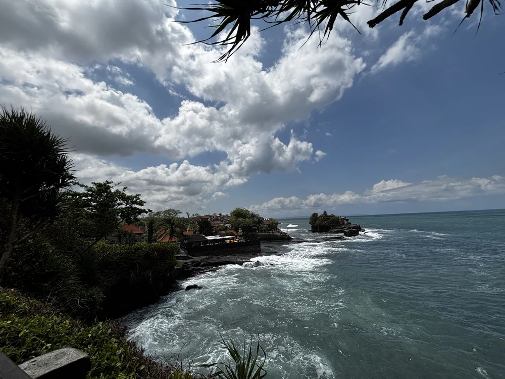 The Tanah Lot temple rock seen in the middle distance from the clifftop walkway, with village rooftops along the shore, white water breaking around the base and the Indian Ocean beyond, Bali