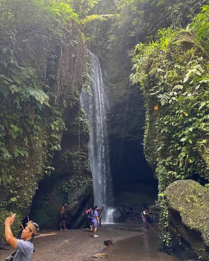 Goa Raja Waterfall: a tall thin drop down a moss-covered canyon wall in Bangli, Bali, with visitors at the base