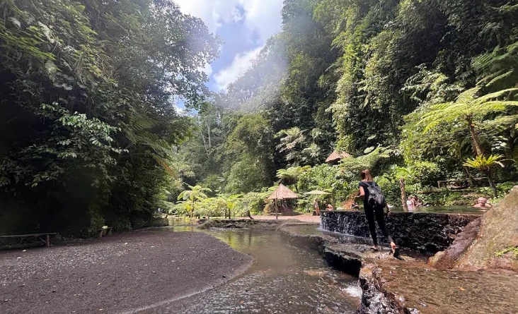 The built stone-rimmed pool below Goa Raja Waterfall with water spilling over its edge, thatched huts and jungle behind
