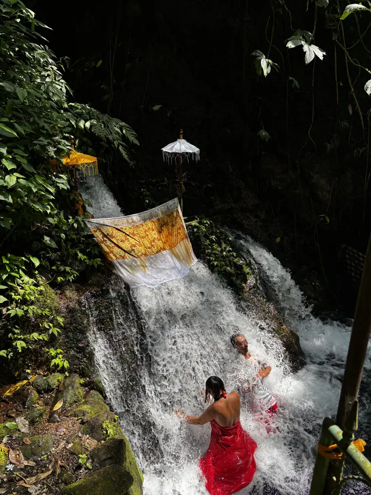 A visitor in a sarong bowing under a cold spring-water spout during a melukat cleansing near Ubud, Bali