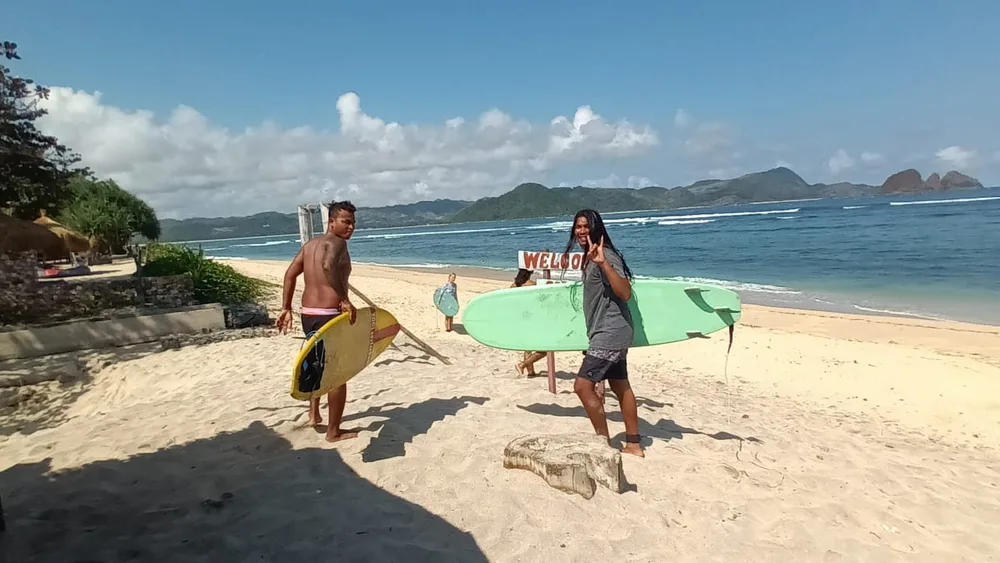 Local surf guides carrying surfboards along a sunny Bali beach with green headlands and turquoise water